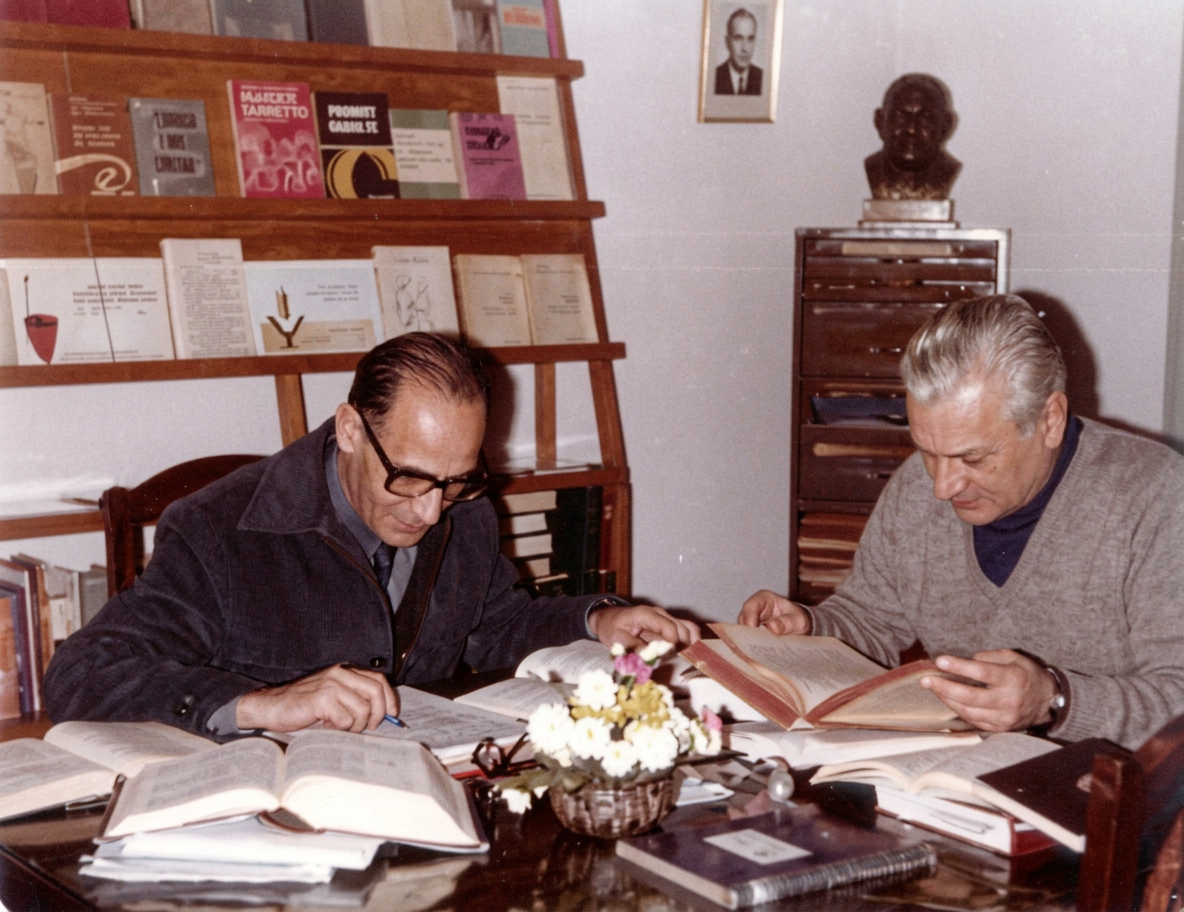 1474459945-300x231-1 Dos hombres sentados en una mesa, revisando libros y documentos, rodeados de estantes llenos de libros. Hay un ramo de flores en la mesa.