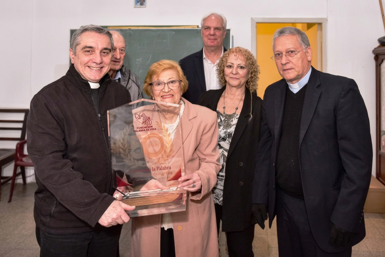 202609261131-163 Un grupo de seis personas, incluido un sacerdote, posando sonrientes en un aula. Una mujer mayor sostiene un premio transparente que dice 'Fundación Palabra en Vida'.